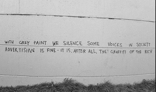 Graffiti on a gray wall. It reads: 'With grey paint we silence some voices in society. Advertising is fine; it is, after all, the graffiti of the rich.'