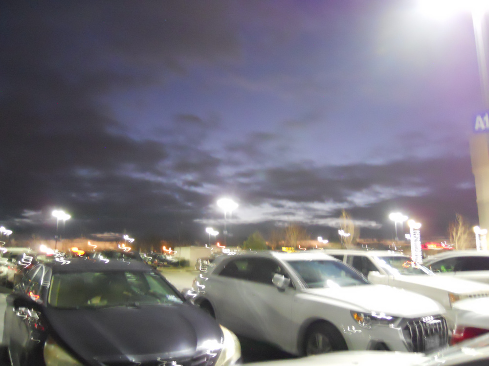 A blurry photo of cars in a parking lot, gleaming under street lamps. Beyond, the sky appears cloudy and purple.