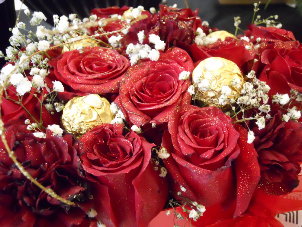 A close-up of an abundant bouquet of rich red roses, their petals glittering with tiny specks of gold dust. Nestled between the roses, there are chocolate truffles in golden wrappers.
