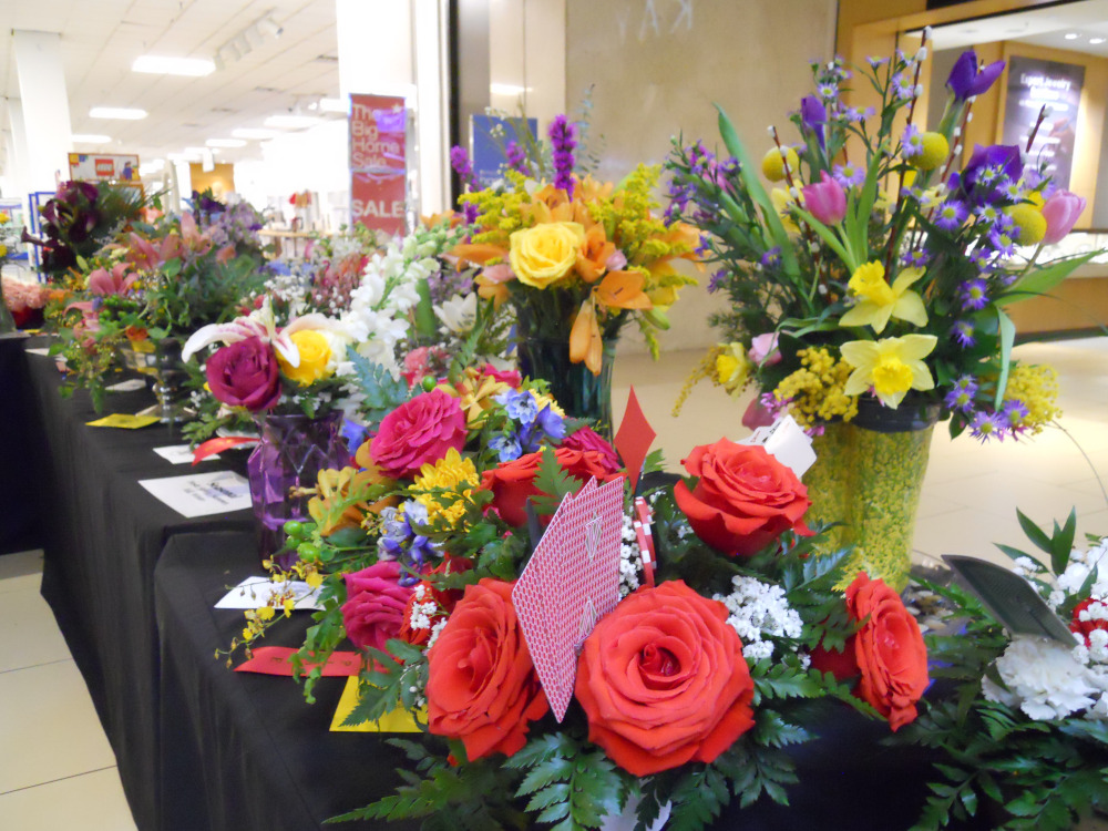 A black table holding around 20 elaborate floral bouquets is set up in the middle of a mall.
