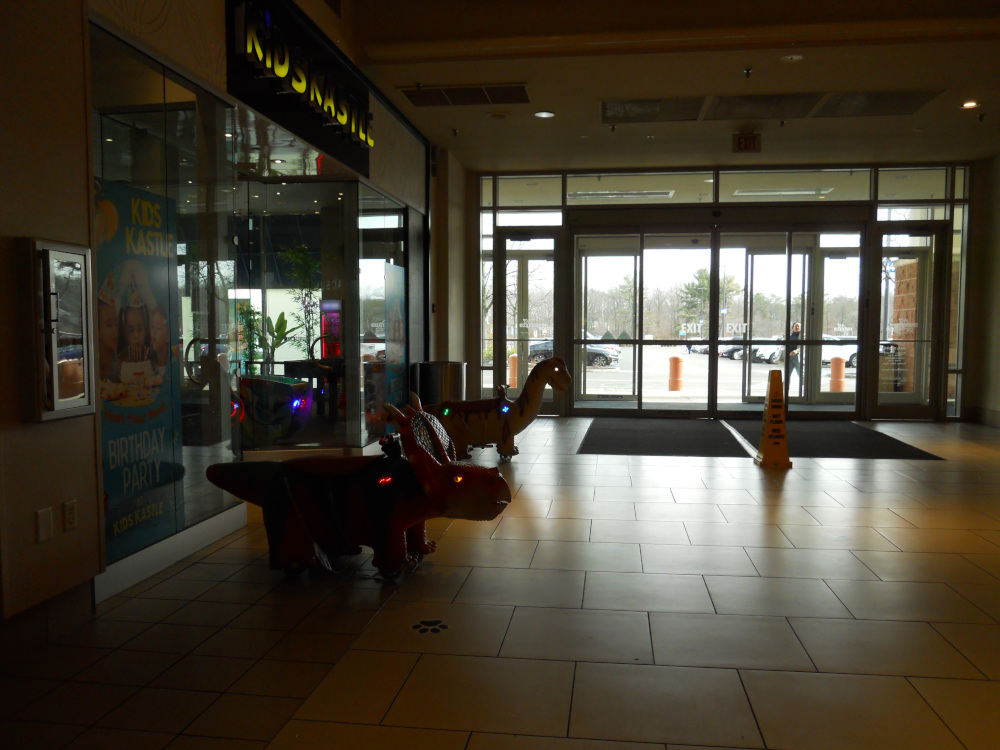 Two animatronic dinosaurs with glowing eyes rest in a dark mall, waiting by a store called Kids Castle.